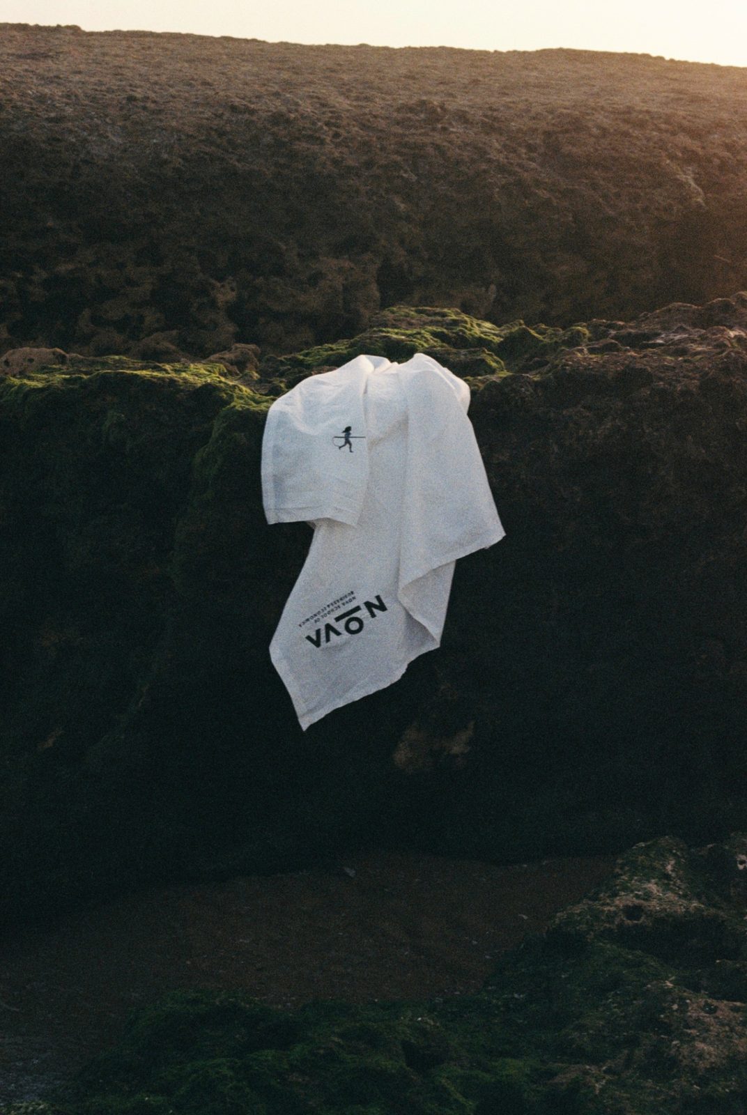 A white printed cloth draped over dark moss-covered coastal rocks, with warm light along the ridge behind.