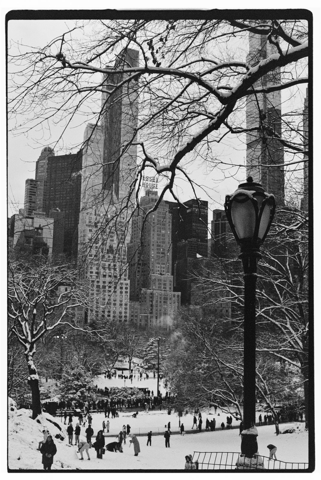 Black-and-white photograph of a snowy park with sledders, a lantern in the foreground and city buildings behind.